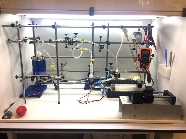 Universal Vaping Machine in a laboratory fume hood with glassware, impinger-style capture, tubing, and instrumentation — a standardized aerosol-capture setup for regulatory chemical analysis